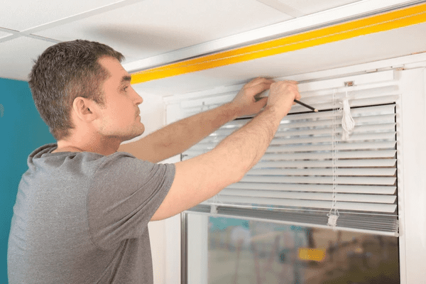 Man repairing white venetian blinds using a screwdriver on a bright window frame.
