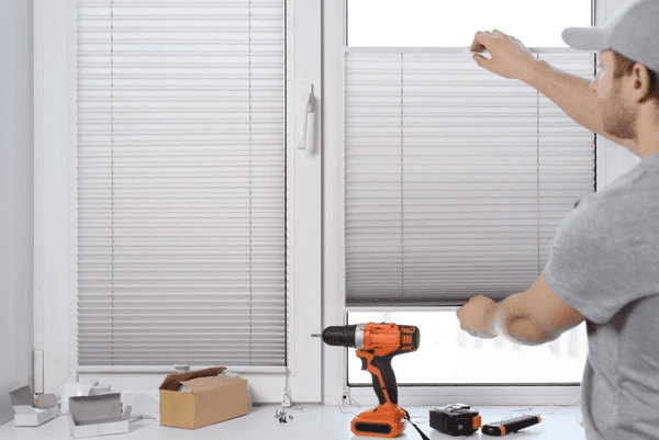 Man installing gray pleated blinds on a window with tools and equipment on the windowsill.