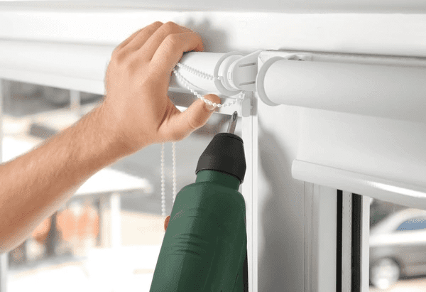 Close-up of a hand repairing white roller blinds with a power drill in a modern Dubai home.