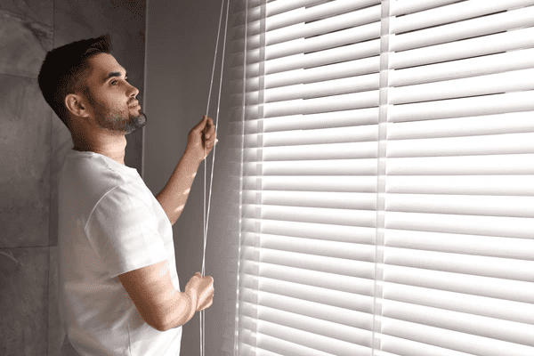 Man in white shirt adjusting sleek white Venetian blinds in a sunlit modern room, perfect for 2025 Dubai interiors that demand elegance and control.