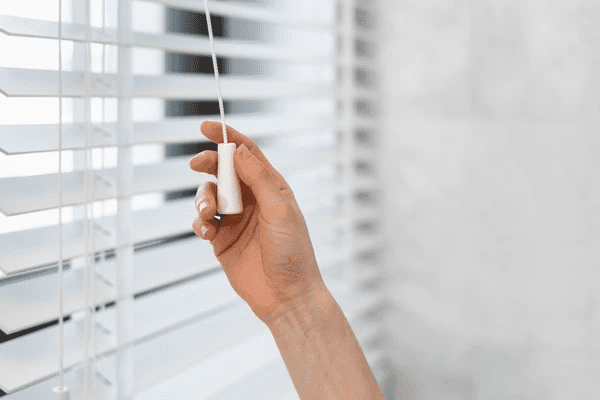 Close-up of a hand adjusting white venetian blinds with a pull cord, set in a modern interior