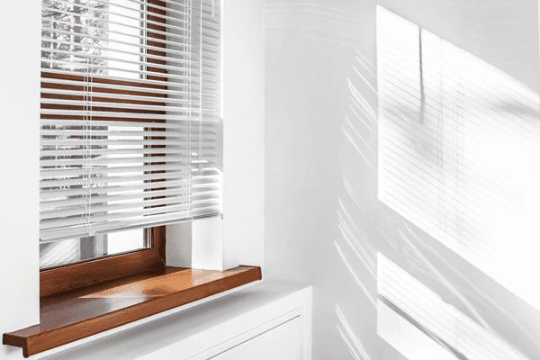 White venetian blinds with sunlight casting shadows on a bright white wall and wooden windowsill in a cozy room