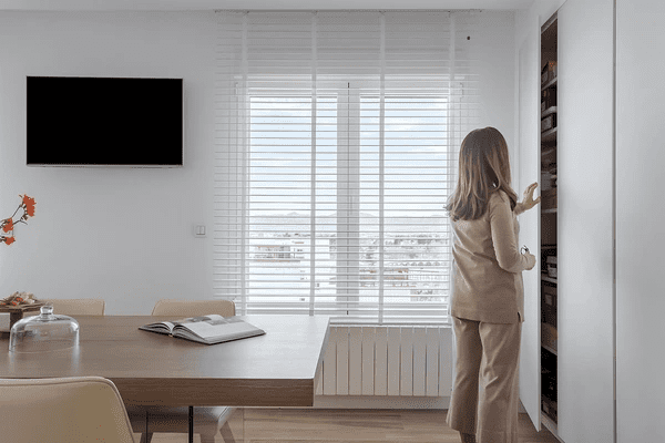 Woman in a beige outfit stands in a bright modern room near white window blinds, accessing a shelf cabinet in a cozy, elegant home interior.
