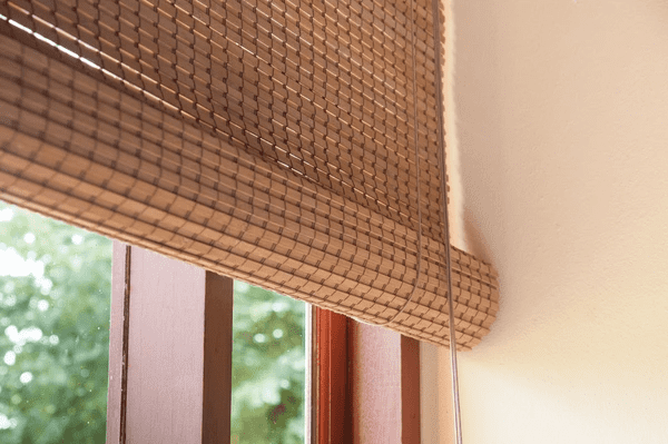 Close-up of eco-friendly wooden blinds rolled up partially, hanging beside a window with a natural outdoor view in the background.