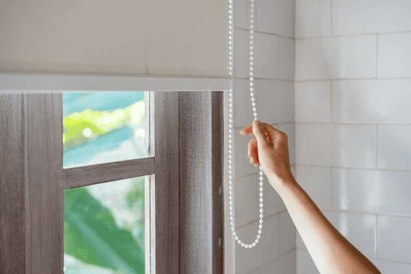A close-up of a hand pulling a white bead cord to adjust modern window blinds in a bright, stylish room.