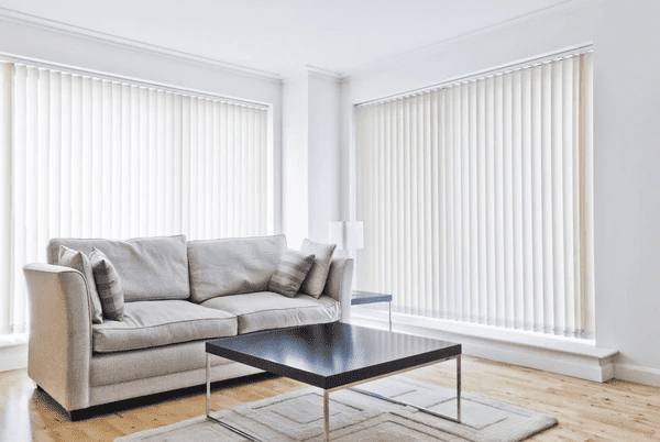 Bright, minimalist living room with large windows covered in elegant cream-colored vertical blinds, soft beige sofa, and a modern black coffee table.