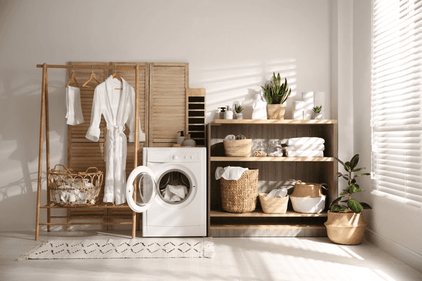 Sunlit laundry area with white washed blinds, modern washing machine, wooden shelving, indoor plants, and cozy textiles.