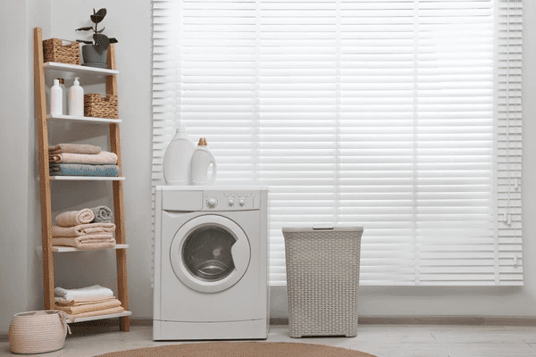 White horizontal blinds in a cozy laundry room setup with a washing machine, detergent bottles, and a wicker basket.