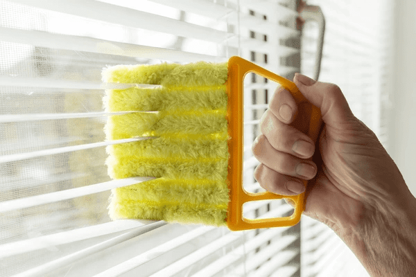A close-up of a hand using a multi-layered microfiber duster to clean white window blinds effortlessly.