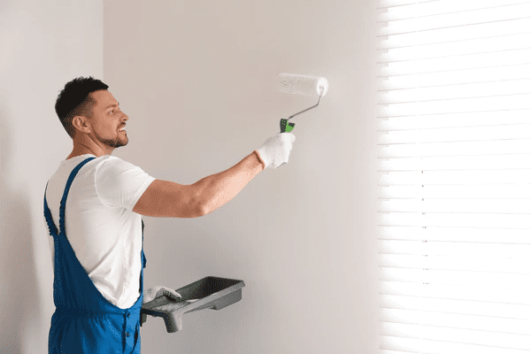 A professional painter in blue overalls carefully applies a fresh coat of paint to elegant white blinds, highlighting the seamless blend of modern aesthetics and luxury.