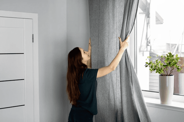 Woman opening sleek grey anti-condensation curtains in a modern Dubai home near a bright window and indoor plant.