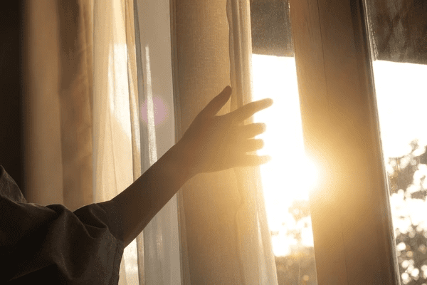 Hand reaching out to adjust climate-friendly soundproof curtains, allowing sunlight to filter through while maintaining privacy.