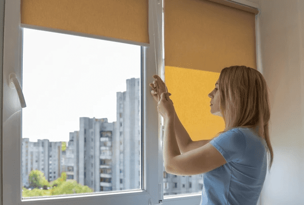 Woman adjusting customized yellow roller blinds in a sleek modern apartment overlooking city buildings in Dubai 2025.