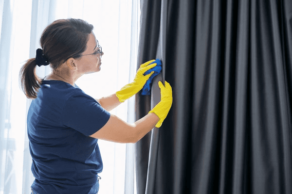 Woman wearing yellow gloves cleaning the Best Blackout Curtains with a blue tool near a bright window.