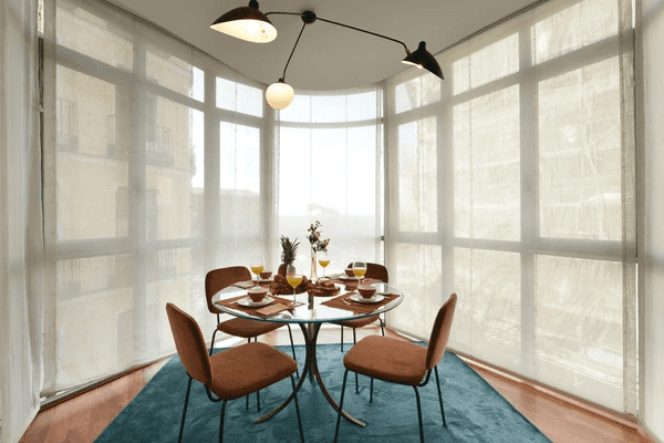 Modern dining area with circular glass table, velvet chairs, and sheer home & furniture blinds covering floor-to-ceiling windows.
