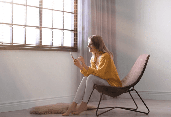 Young woman using a smartphone near a sunlit window with smart sheer curtains in a sleek, minimalist room.