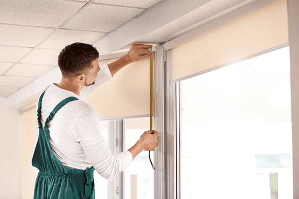 Handyman in green overalls using a tape measure to install roller blinds on large windows in a modern rental property.