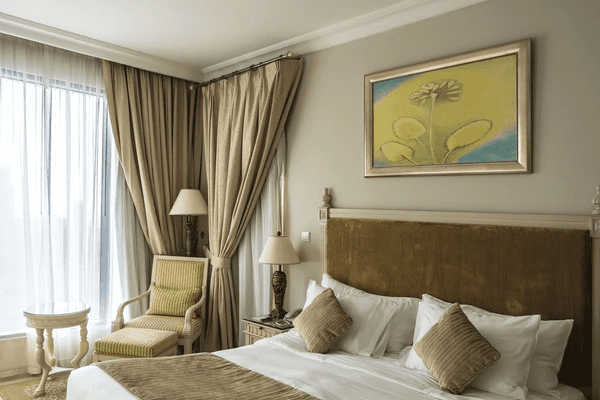 Elegant hotel-style bedroom in Dubai with golden curtains, beige headboard, classic furniture, and framed artwork above the bed.