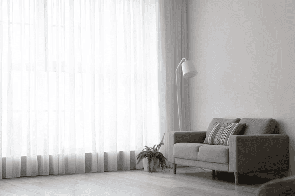 Bright living room with floor-to-ceiling white sheer curtains, a modern grey sofa, a floor lamp, and a potted plant.