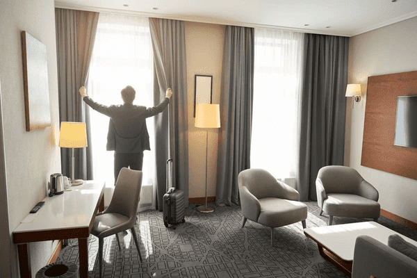 Man in travel attire opening thick blackout curtains in a modern hotel-style Dubai living room.
