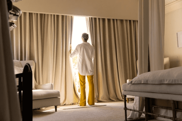 Senior woman standing by a large window framed with elegant full-length beige track curtains in a cozy bedroom.