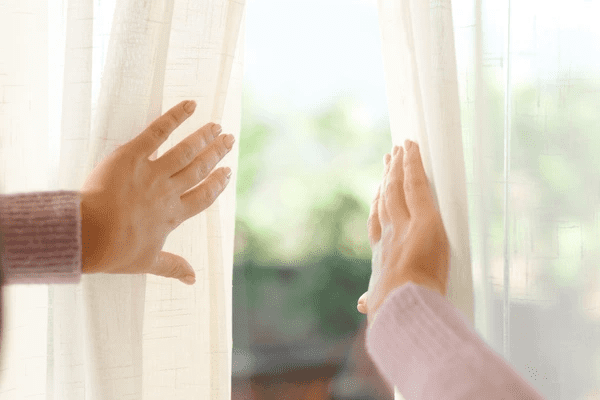A close-up shot of hands opening soft, light-colored soundproof curtains to reveal a serene outdoor view. The soft fabric of the curtains adds a peaceful, stylish atmosphere to the room.


