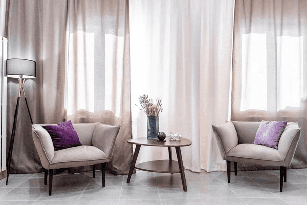 A cozy living room featuring beige and white soundproof & acoustic curtains, two plush armchairs with purple cushions, and a decorative table with a vase of dried flowers. The soft natural light creates a peaceful, stylish atmosphere.

