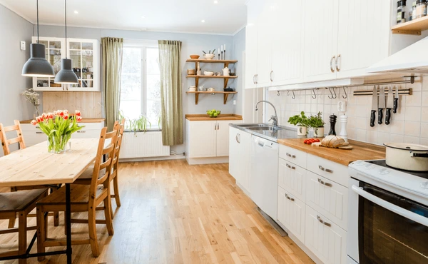 Scandinavian-style kitchen in Dubai with wooden flooring, white cabinetry, and short olive-green curtains over a large window.

