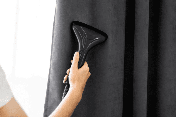 A close-up of a woman using a steamer to remove wrinkles from a dark soundproof curtain. The fabric is being carefully steamed to maintain its sleek, modern look and functionality.

