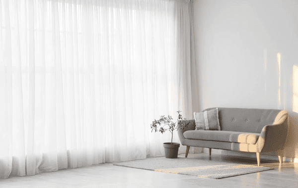 Elegant white sheer curtains hanging in a bright Dubai living room with soft natural light, grey furniture, and modern décor.

