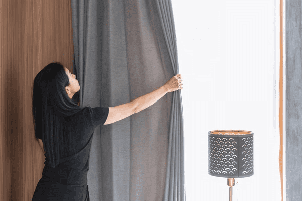 A woman pulling back a sleek gray soundproof curtain, allowing natural light to flood the room. The modern, stylish curtains are complemented by a chic lamp, creating a peaceful atmosphere in the room.

