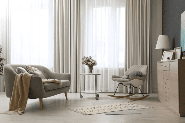 A modern living room featuring beige soundproof & acoustic curtains, a cozy sofa, and a rocking chair. The space includes soft lighting, a small table with flowers, and elegant décor elements.

