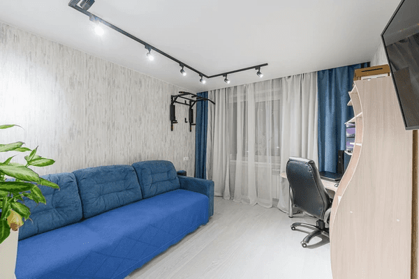 Modern small room in Dubai with blue track curtains, a blue sofa bed, desk setup, and ceiling track lighting.

