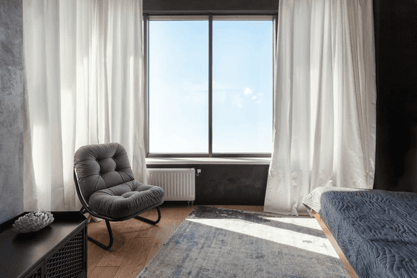 A cozy bedroom featuring light, soundproof & acoustic curtains with a large window offering a bright outdoor view. The space includes a comfortable gray chair, a soft rug, and modern minimalist furniture.

