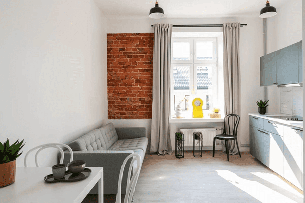 A cozy, modern loft apartment with gray soundproof curtains framing a window. The room features an industrial brick wall, minimal furniture, and natural light filling the space.

