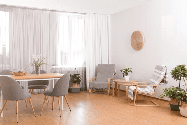 A bright and airy living room with stylish soundproof & acoustic curtains, a dining area with gray chairs, and minimalist furniture. Plants are placed throughout the room, creating a natural, calm ambiance.

