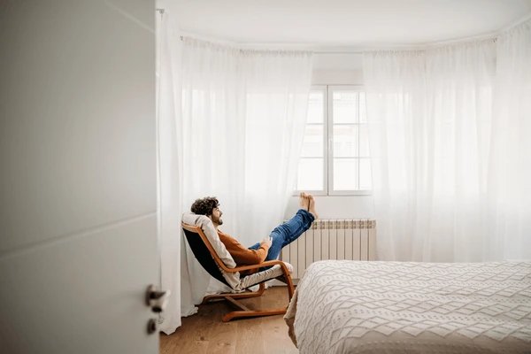 Cozy bedroom in Dubai with minimalist decor and elegant white sheer curtains, featuring a man relaxing on a lounge chair.