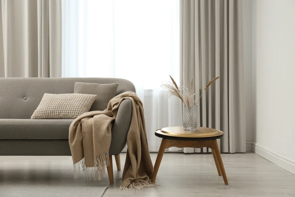 Modern living room with soft beige curtains, grey sofa, textured cushions, and a wooden coffee table in Dubai.