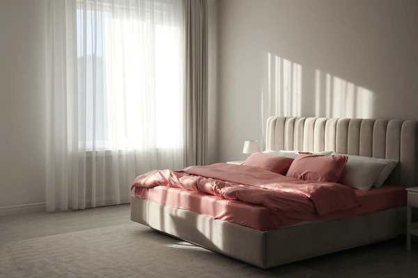 Sheer white linen curtains filtering natural sunlight in a minimal bedroom with a soft pink bed setup.