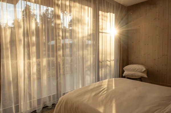 Bedroom in Dubai with sheer floor-to-ceiling curtains, sunlight streaming through large glass windows, and a cozy minimalistic setup.
