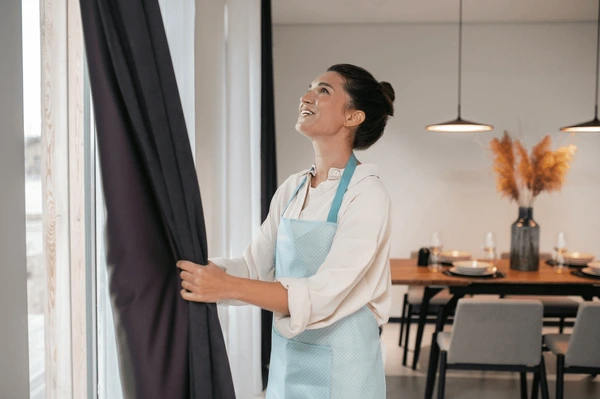 Woman adjusting fire-resistant curtains in a stylish Dubai dining room with warm lighting and modern décor.