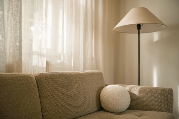 Minimalist living room with beige linen curtains, textured sofa, round cushion, and floor lamp in warm sunlight.

