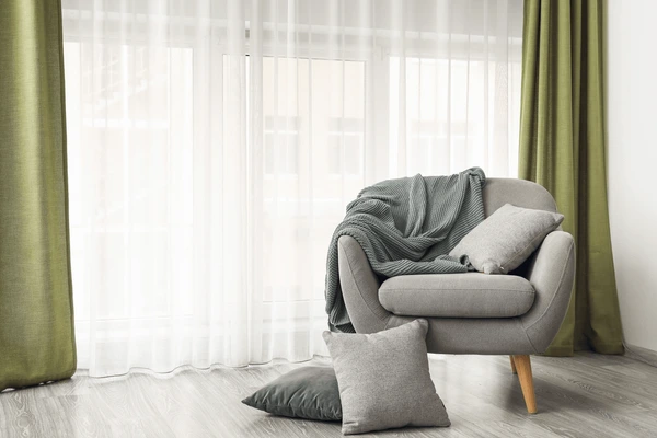 Cozy armchair with cushions and a throw blanket beside layered green and sheer white curtains, adding elegance and comfort to a Dubai home interior.