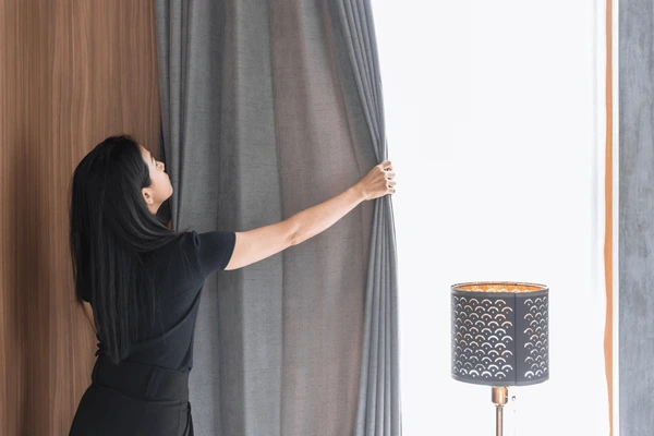 Woman pulling back gray summer curtains in a sunlit Dubai room, revealing a bright, sleek interior with a modern lamp.