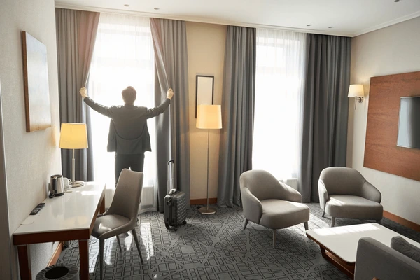 Business traveler opening grey blackout curtains in a modern, well-lit hotel room with elegant furniture and neutral decor.