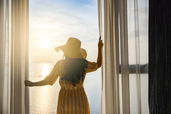 A woman in a yellow striped dress opens sheer white curtains to a bright summer sunrise over calm waters in a Dubai home.