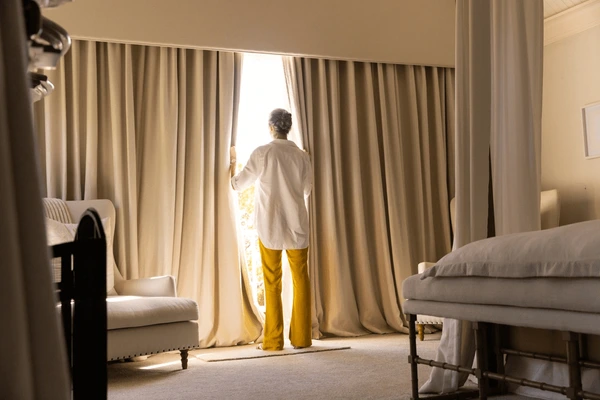 A person standing by large beige curtains in a sunlit bedroom, creating a cozy, modern, and elegant ambiance.