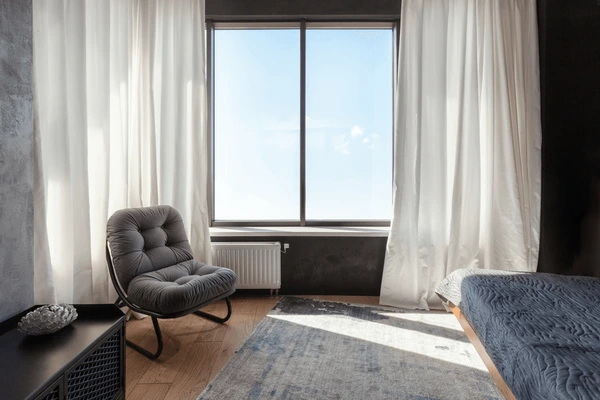 Best normal sized short curtains in Dubai 2025 with elegant white drapes framing a modern bedroom featuring a cushioned chair, stylish bed, and soft rug.