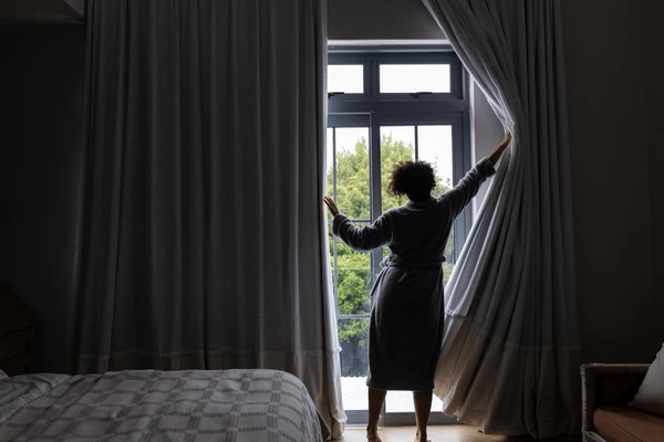 Best Grey Wall Curtains in Dubai 2025 showing a woman in a bathrobe opening luxurious grey curtains in a modern bedroom with natural light.