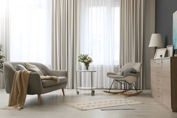 Elegant living room with grey wall curtains, soft grey sofa, rocking chair, wooden cabinet, and natural light in Dubai 2025.